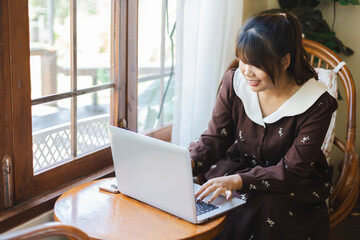 Asian woman is happily working on her laptop as digital nomad enjoying remote work by window in...