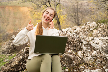 Young woman working remotely with laptop, talking on smartphone in nature park, looking at camera,...