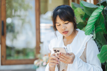 Asian woman is working remotely as digital nomad using her smartphone outdoors near green plants...