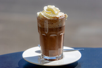 Selective focus of hot chocolate in a glass served with white whipping cream on the top, Warm dark brown hot drink on wooden table outdoor terrace under sunlight.
