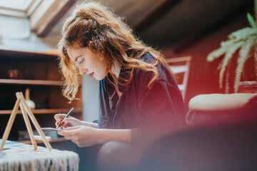 A young woman with curly hair is depicted intently working on an art project in her cozy living space, expressing creativity and focus while drawing or painting on a surface.