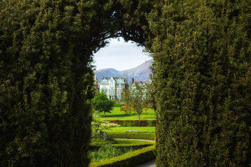 Manicured garden with topiary arch framing a view of a traditional English manor house. Landscape design with trimmed hedges, lawns, distant hills. Luxury real estate, countryside tourism, travel