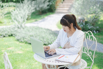 Asian woman is working remotely as digital nomad using laptop outdoors in peaceful garden setting...