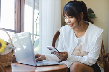 Asian woman digital nomad remote working happily on laptop and smartphone at home near window...