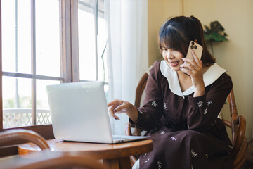 Asian woman working remotely as digital nomad smiles while using laptop and talking on phone in...