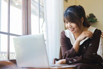 Asian woman is happily working on her laptop as digital nomad enjoying remote work by window in...