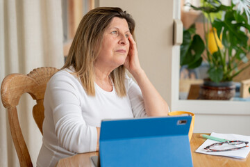Thoughtful senior woman using a digital tablet at home, contemplating the next steps in her online...