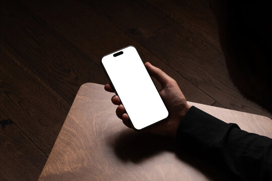 A person holding smartphone mockup on a wooden table in a dark cozy room with wooden floor. Isolated blank surface to place your design.