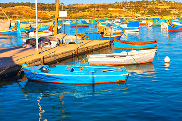 Serene harbor scene with numerous small boats, many painted in bright colors, docked along a wooden pier. In background, a hilly landscape with sparse vegetation stretches under a clear sky