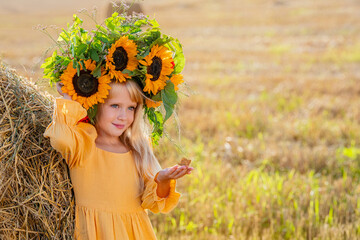 A young girl in a bright crown of sunflowers and a yellow dress leaning against a haystack in a...