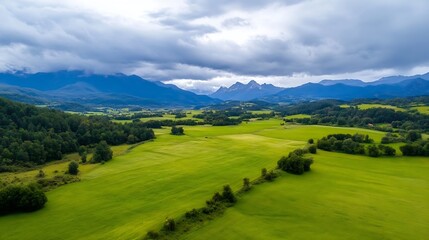 Obraz premium Aerial view of a lush green valley nestled amongst majestic mountains under a dramatic cloudy sky. The vibrant green fields contrast beautifully with the dark blue mountain peaks, crea : Generative AI