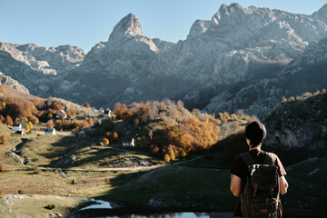 Naklejka premium Female traveler with backpack looking rocky peaks of Montenegro near Bukumirsko Lake. Golden autumn trees remote mountain village and peaceful wilderness in the Balkans. Rear view.