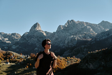 Naklejka premium Female solo hiker with a backpack admires the majestic peaks of Montenegro in warm autumn light. The village below Bukumirsko Lake glows with vibrant fall colors in this cinematic mountain landscape.