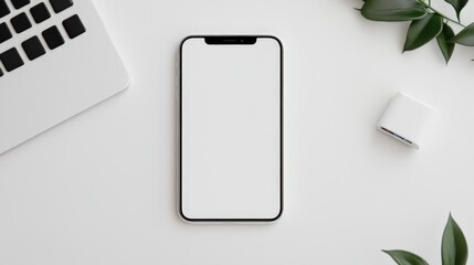 A modern smartphone with a blank screen lies on a white desk near a laptop, earbuds case, and green leaves.