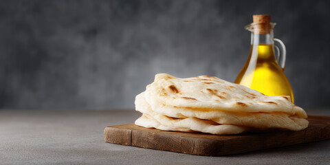 Flatbread is stacked on a wooden board with a glass bottle of oil beside it. Rustic cooking concept