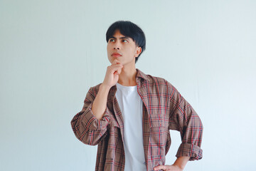 Asian man holding his chin and looking up sideways with a thinking expression, isolated on a plain...