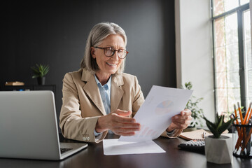 Smiling mature business woman entrepreneur working in office checking legal document account invoice in office. Businesswoman of middle age manager executive or lawyer using laptop computer at work.