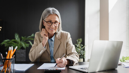 Happy mature mid aged business woman professional in her 50s writing notes finance report overview, lawyer attorney checking document, working on laptop computer device sitting at desk in office.