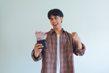 Portrait of happy young Asian man holding wallet full of cash and showing victory gesture, isolated...