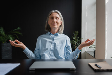 Calm Caucasian mature female sits in relaxed yoga position at desk in front of laptop with eyes...