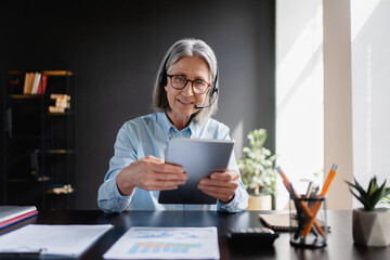 Busy middle aged female support service operator talking to customer sitting at desk. Happy mature...