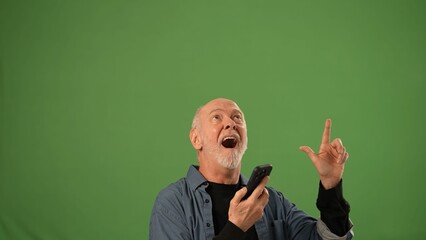 A mature man shows excitement while pointing upward and holding a smartphone, with a solid green background creating an ideal space for product announcements.