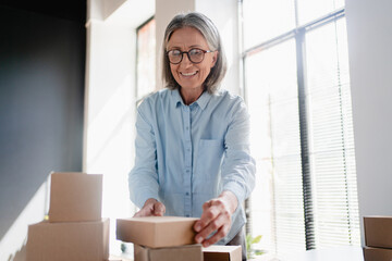 Older mature female online store small business owner worker packing package post drop shipping ecommerce retail order in box preparing delivery parcel on table in warehouse. Dropshipping service.
