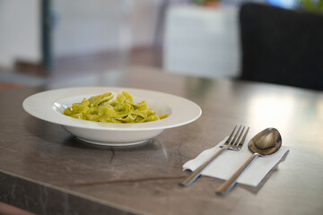 Fettuccine pasta with pesto sauce, basil, and parmesan cheese on a white plate, a spoon, and a fork is shown on the top view of a table.