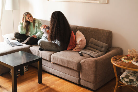 Two women are relaxing on a couch. The blonde caucasian female is smiling at the Arab female with curly dark hair. They are enjoying time together in a cozy living room.