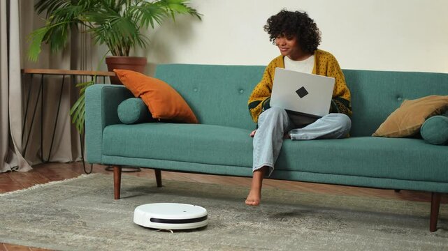 Robotic vacuum cleaner cleaning room while woman working with laptop on sofa. Woman sitting on couch using laptop, robot vacuum cleans floor. Smart home automation modern technology in daily life