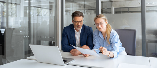 Team of partners mature man and young woman discussing financial banking document report sitting at table in office. Two colleagues professional business people working together on project. Copy space