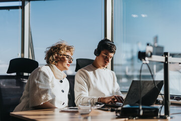 Two business employees collaborate at a modern office desk, one wearing headphones. They focus...