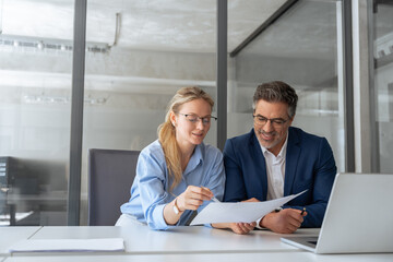 Team of diverse partners mature man and young woman discussing financial document using computer sitting at table in office. Two colleagues of professional business people working together on project