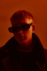 Fototapeta premium Close-up portrait of young stylish guy in trendy sunglasses and black coat, posing against red studio background in neon light. Concept of bold fashion, style, youth culture, self-expression