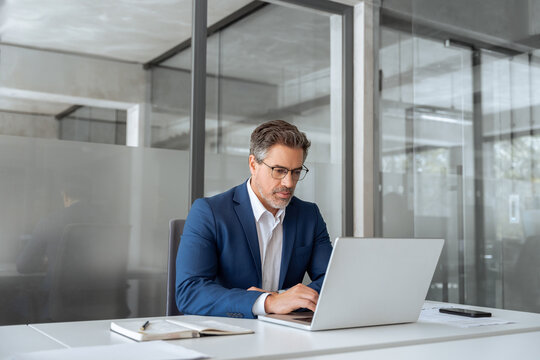 Mature indian ceo business man in suit using pc laptop for trading, looking focused at screen. Middle age latin male entrepreneur businessman working on financial work at desk in office. Copy space