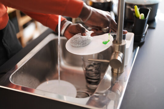 A person is washing dishes in a stainless steel sink. They are using a metal scrubber and green dish soap to clean a white plate.