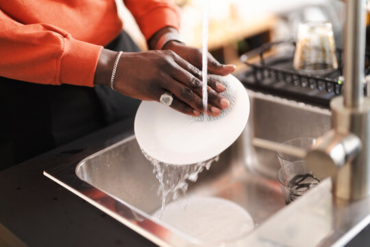 A person wearing an orange sweater washes a white plate with a sponge under running water in a stainless steel sink. The individual is wearing jewelry.