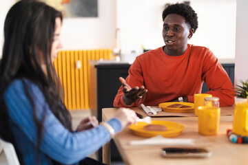 A young couple is having breakfast at home. They are sitting at the table, eating pancakes and talking.