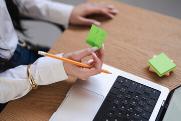 A woman holds a sticky note reminding her of a meeting at 11:00 AM. She is working at a laptop with a pencil in her hand, ready to take notes.