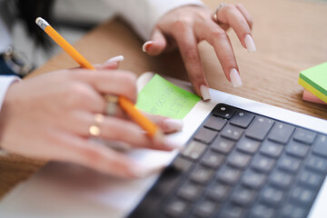 A woman is writing a reminder on a sticky note with a pencil. The note is placed on a laptop, and she is organizing her schedule.