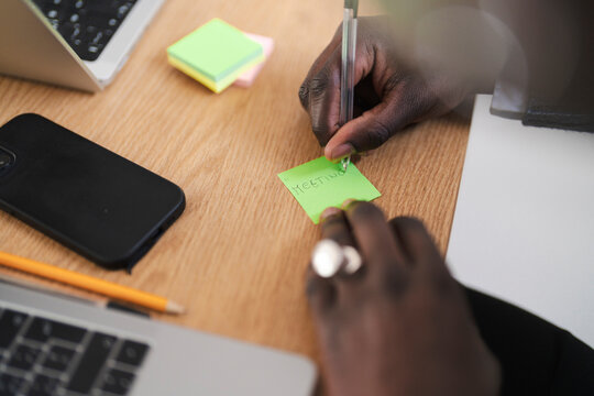 A person writes "meeting" on a sticky note at their desk. They are using a pen and have a laptop and phone nearby.