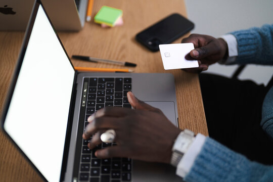 A person is using a laptop and holding a credit card. They are shopping online or paying bills. The scene is set on a wooden desk with office supplies.