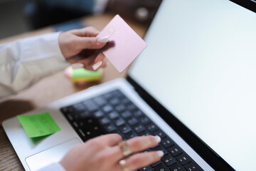 A woman is shopping online. She's holding a pink credit card and typing on her laptop to complete the purchase.