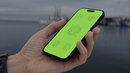 GREEN SCREEN CHROMA KEY Hand holding smartphone with green screen at a harbor on an overcast day, ferry in the background, ideal for customizable content related to travel or ticketing