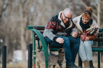 Smiling couple sitting on an outdoor bench while drinking beverages and interacting with a cat.