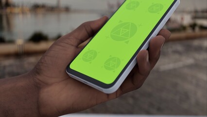 GREEN SCREEN CHROMA KEY person holding a smartphone with green screen near a marina with yachts and high-rise buildings in the background during sunset