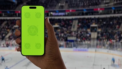 GREEN SCREEN CHROMA KEY A hand holding a phone with a green screen at a hockey game in an indoor stadium, ideal for showcasing live sports or betting apps