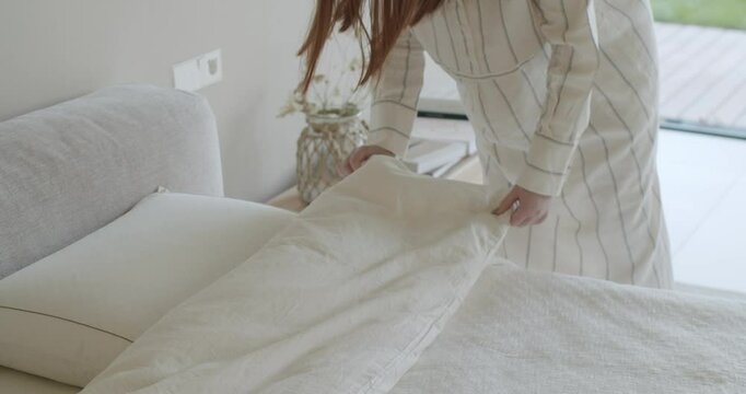 Woman in pajamas makes the bed in the bedroom. She covers the bed with a blanket. Daily routine, morning, Woman Making Bed in Cozy Bedroom, Morning Routine and Home Comfort