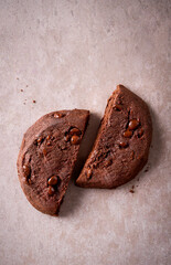Top view of a broken chocolate cookie with visible chips on a beige stone surface. Minimal styling and soft light. Perfect for bakery ads, food blogs, packaging or branding.