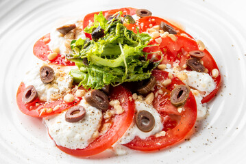 Fresh salad with tomatoes, creamy cheese, black olives, pine nuts, and lettuce on a white plate, vibrant and appetizing, perfect for healthy Mediterranean cuisine.